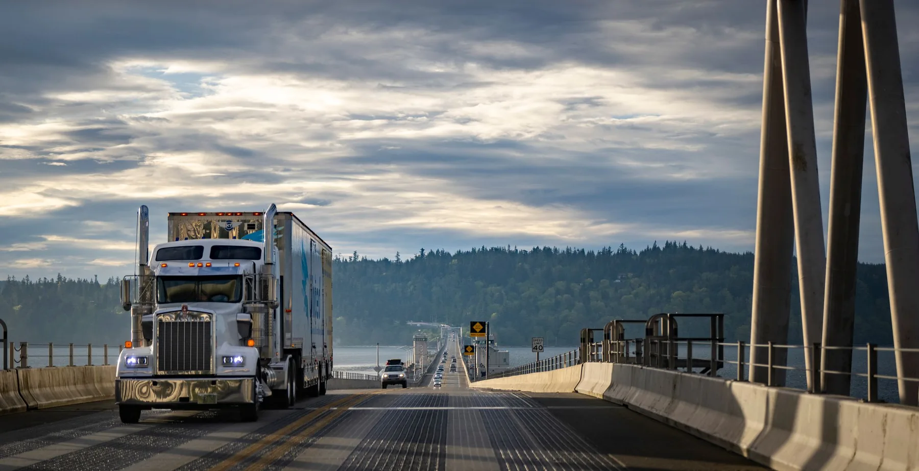 Silver conventional semi-truck crossing a bridge beneath a cloudy sky.
