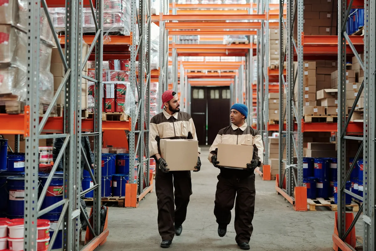 Two workers carrying cardboard boxes through a warehouse aisle.
